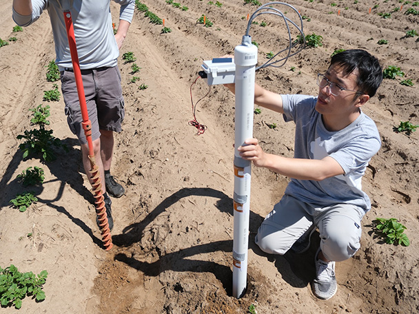 A man pushes a tubular piece of equipment into the ground in a farm field as another person watches.