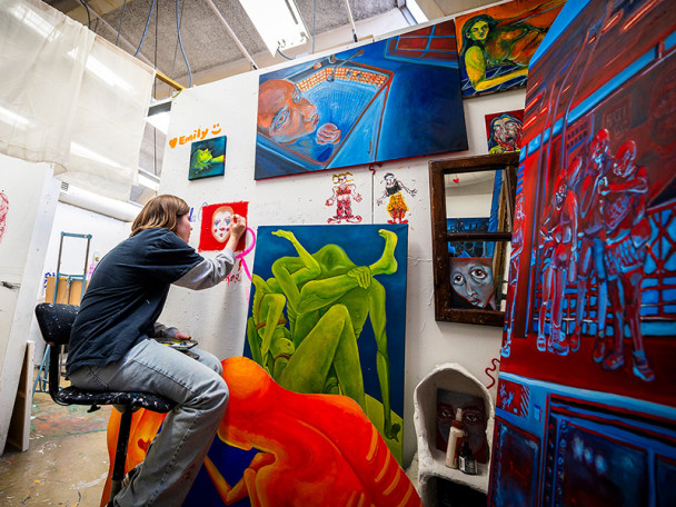 A woman carefully paints a face on a wall in an art studio; the wall is hung with several other colorful paintings.