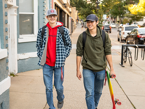 Two young men walk side by side in downtown Madison, wearing backpacks. One carries a skateboard.