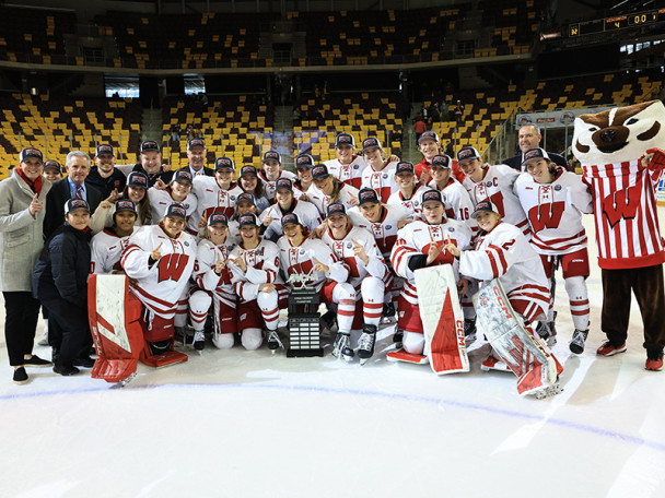 The UW Women's hockey team in red and white uniforms poses for a group photo on the ice with a trophy, surrounded by coaches and Bucky Badger.