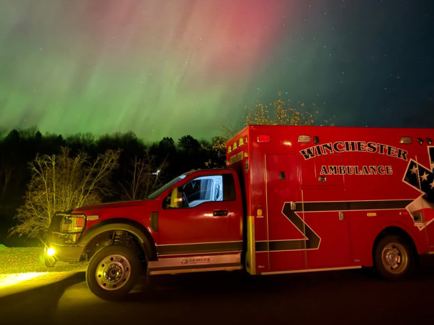 A red ambulance with the words "Winchester Ambulance" on the side of the road at night with Northern Lights in the background