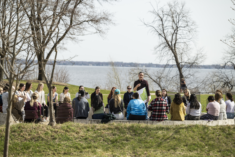 Man speaking to a group of students in front of a lake.